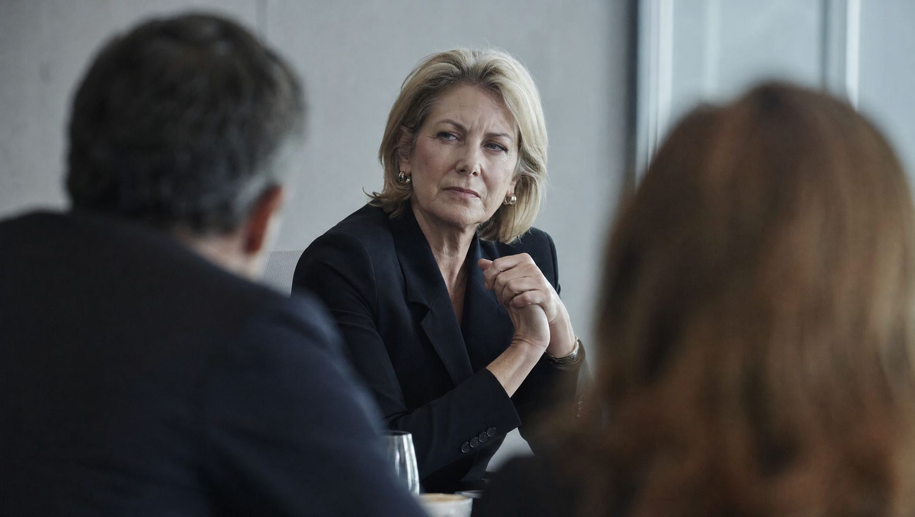 A senior executive listening with a sceptical expression across a boardroom table, framed between two colleagues.