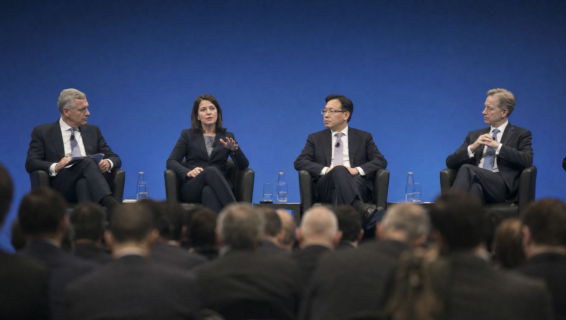 Four executives on a panel at a business forum, one woman speaking, seen over the heads of the audience.