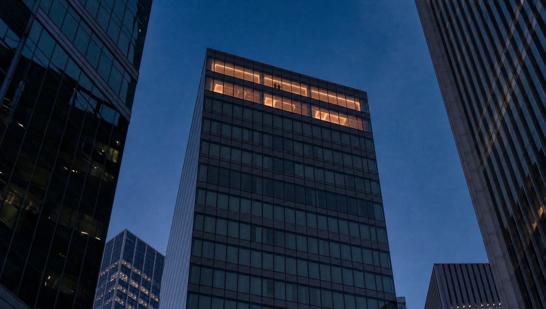 A glass office tower at dusk with only its top floors lit, seen from the street between neighbouring towers.