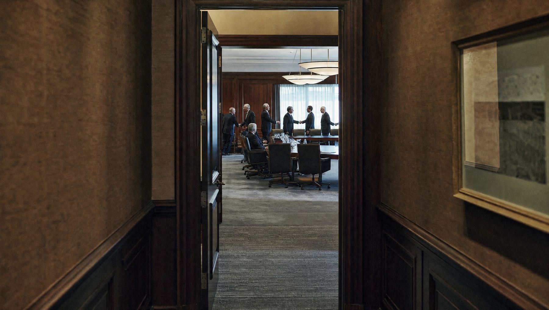 Two leadership teams meeting in a wood-panelled boardroom, seen from a dark corridor through an open door.